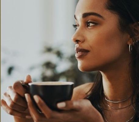 Dark skinned woman holding a cup of tea or coffee looking dreamily towards a window