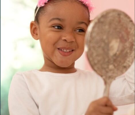 Young African American girl looking at her reflection in a hand mirror and smiling. You cannot see her image, only the back of the hand mirror.
