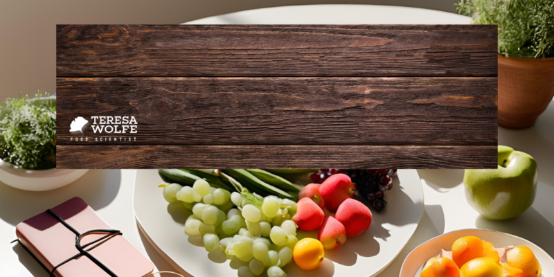 A beautifully arranged plate of fresh fruits, including grapes, apples, and citrus, sits on a white table surrounded by greenery and a notebook. A rustic wooden cutting board with the words 'Holistic Nutrition vs. Dieting—A New Perspective' in white font is placed over the image, symbolizing the contrast between restrictive dieting and a balanced, nourishing approach to food.